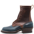 A rugged ankle boot made of dark brown and navy blue leather, featuring metal eyelets, a stacked dark brown heel, and a pull loop at the back, shown against a plain white background.