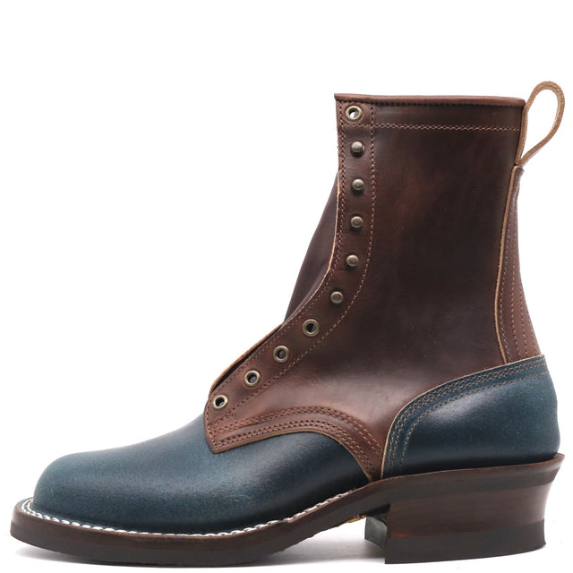 A rugged ankle boot made of dark brown and navy blue leather, featuring metal eyelets, a stacked dark brown heel, and a pull loop at the back, shown against a plain white background.