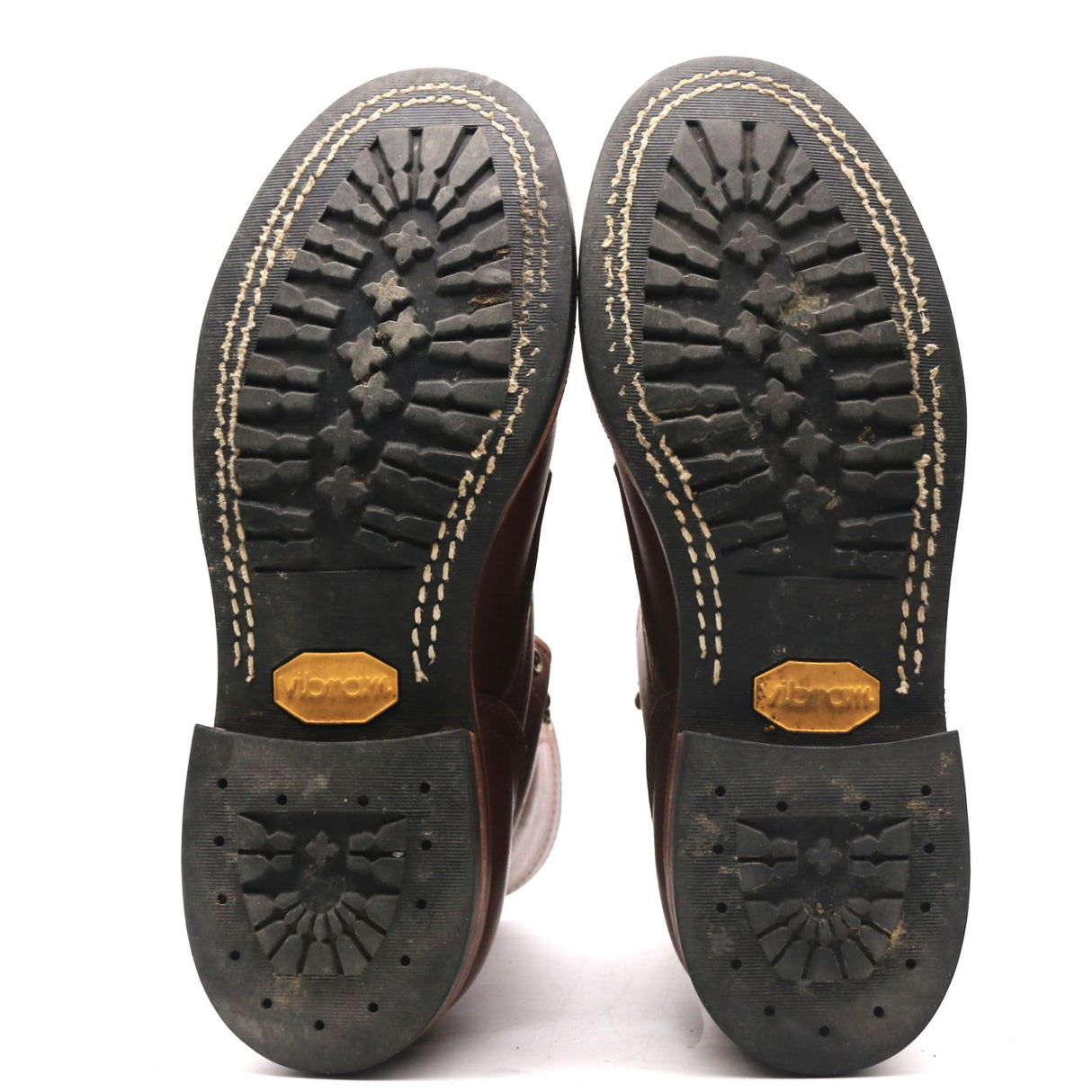 A pair of boots shown from the bottom, displaying the black rubber soles with deep tread patterns, beige contrast stitching, and gold-colored Vibram logos on the heel.