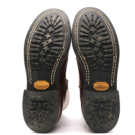 A pair of boots shown from the bottom, displaying the black rubber soles with deep tread patterns, beige contrast stitching, and gold-colored Vibram logos on the heel.