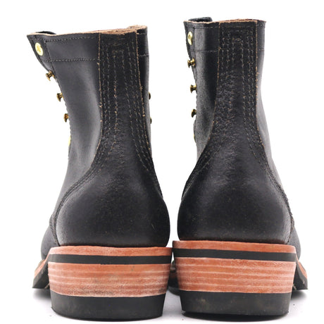 A pair of black leather lace-up boots shown from the back, featuring brass grommets and a stacked brown and black sole.