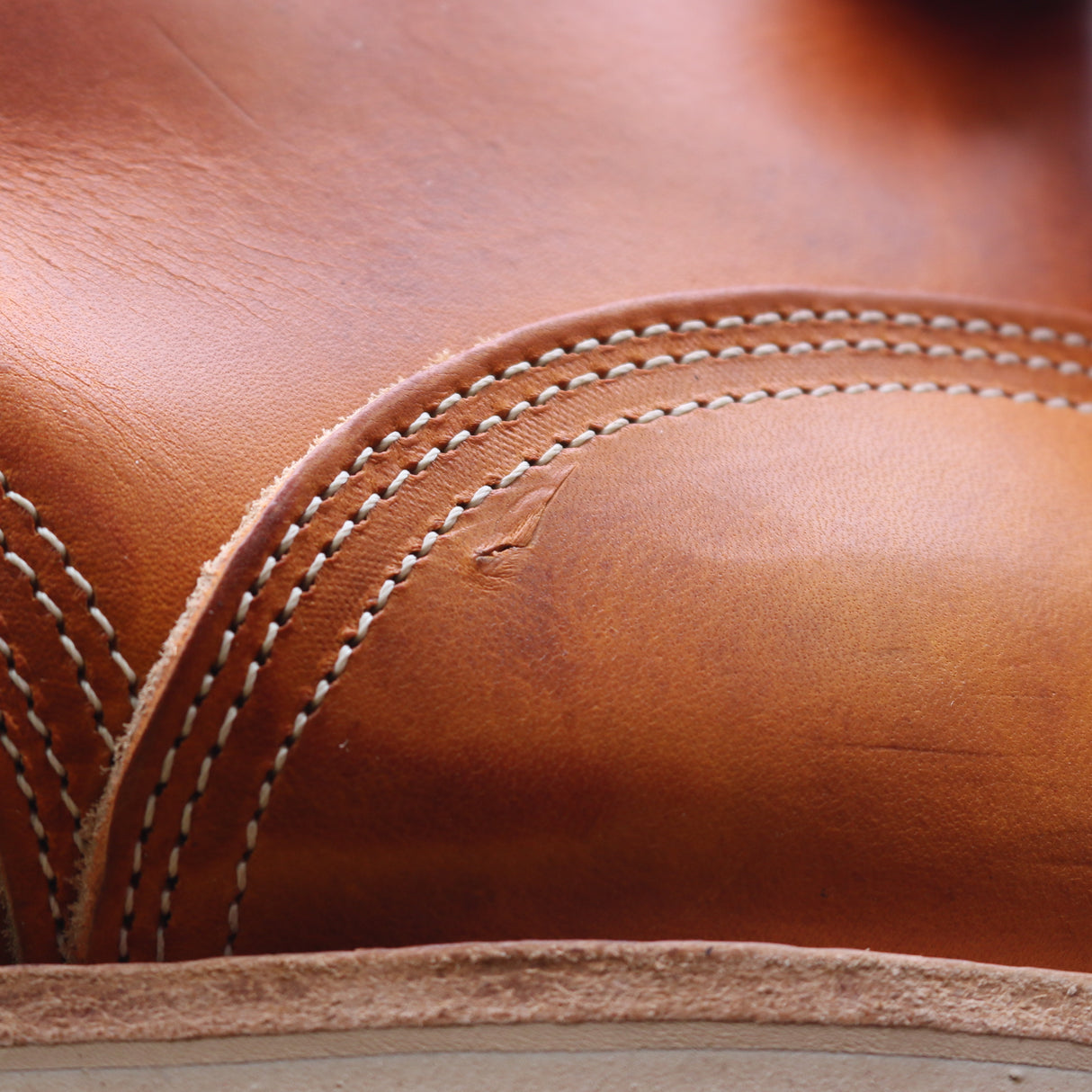 Close-up of a warm tan leather item with white contrast stitching and a small visible scratch or scuff on the surface.
