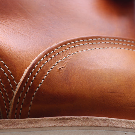 Close-up of a warm tan leather item with white contrast stitching and a small visible scratch or scuff on the surface.