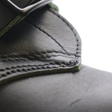 Close-up detail of a black leather strap or belt with visible stitching and a metal buckle, set against a bright white background.