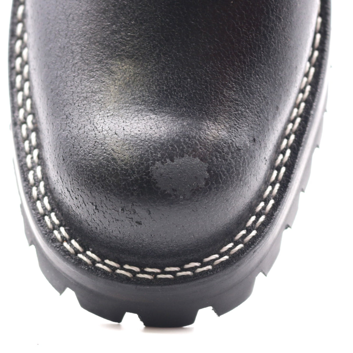 Close-up of a black leather boot toe showing white contrast stitching and a black treaded sole on a white background.