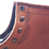 Close-up of a brown leather boot corner showing stitched edge, metal grommets, and rivets against a bright white background.