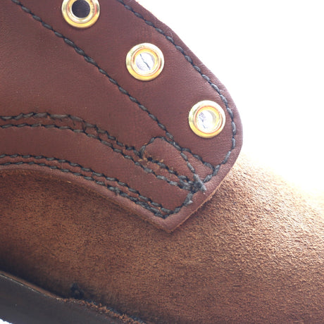 Close-up detail of a brown leather shoe or boot showing a stitched overlay panel with brass grommets and a suede-like brown footbed.