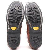 A pair of brown leather boots shown from the sole, displaying the black rubber tread with white stitching and yellow Vibram logos on a white background.
