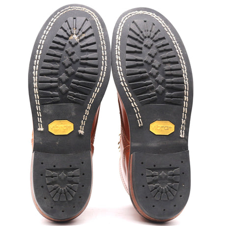 A pair of brown leather boots shown from the sole, displaying the black rubber tread with white stitching and yellow Vibram logos on a white background.