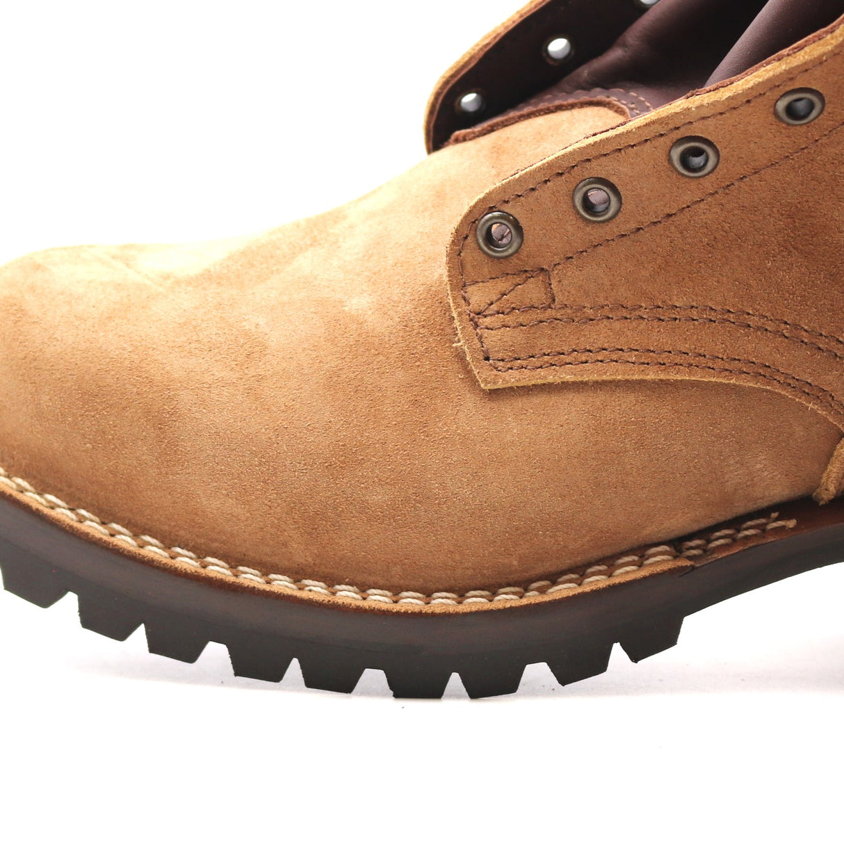 Close-up of a tan suede work boot showing the side profile with metal eyelets, contrast stitching, and a dark lug sole on a white background.