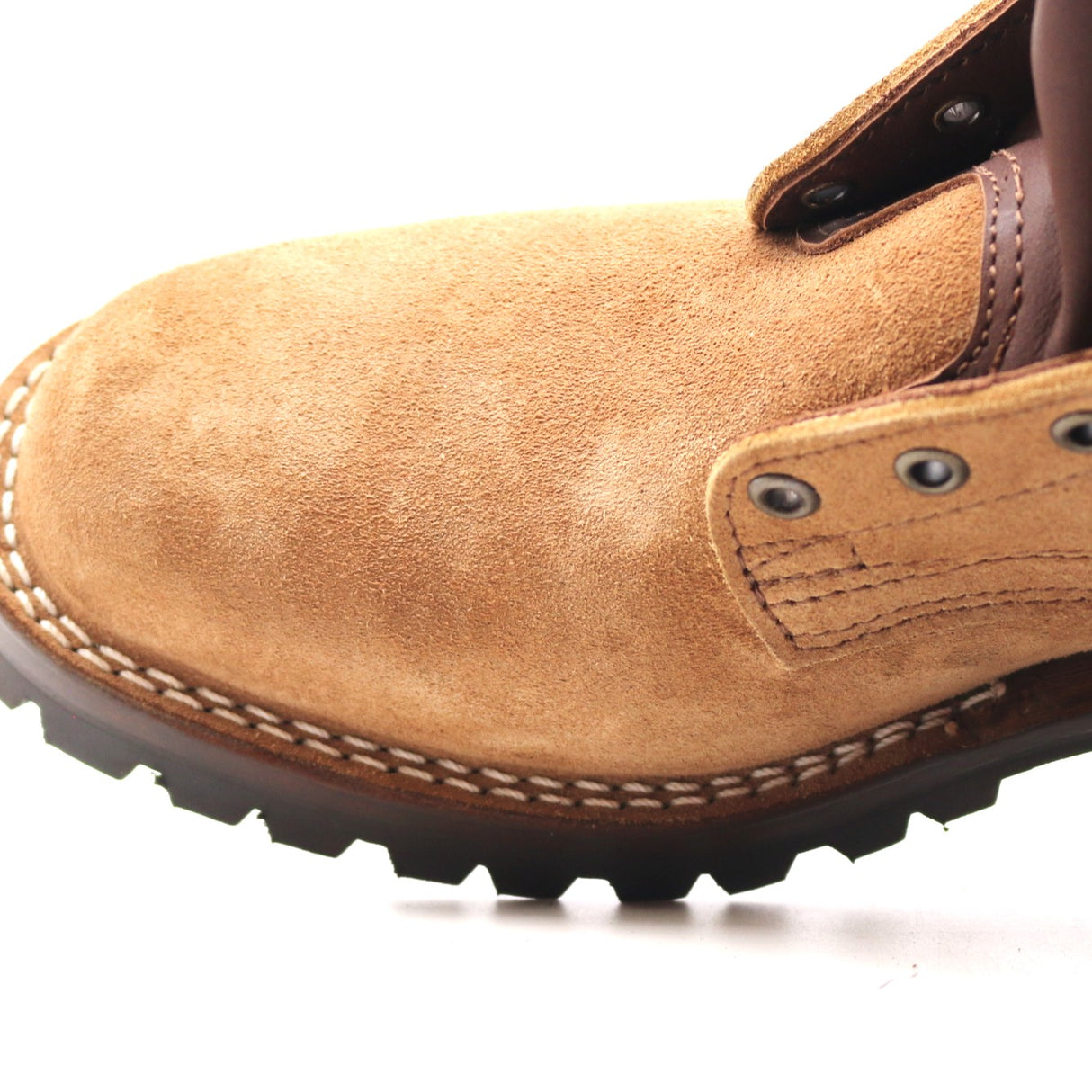 Close-up of a tan suede work boot with dark brown laces, metal eyelets, and a black lug sole on a white background.