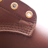 Close-up of a brown leather shoe or boot corner with visible stitching, raw edge, and two brass grommets on a white background.