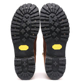 A pair of brown boots shown from the bottom, displaying their black Vibram soles with yellow Vibram logos and metal spikes.