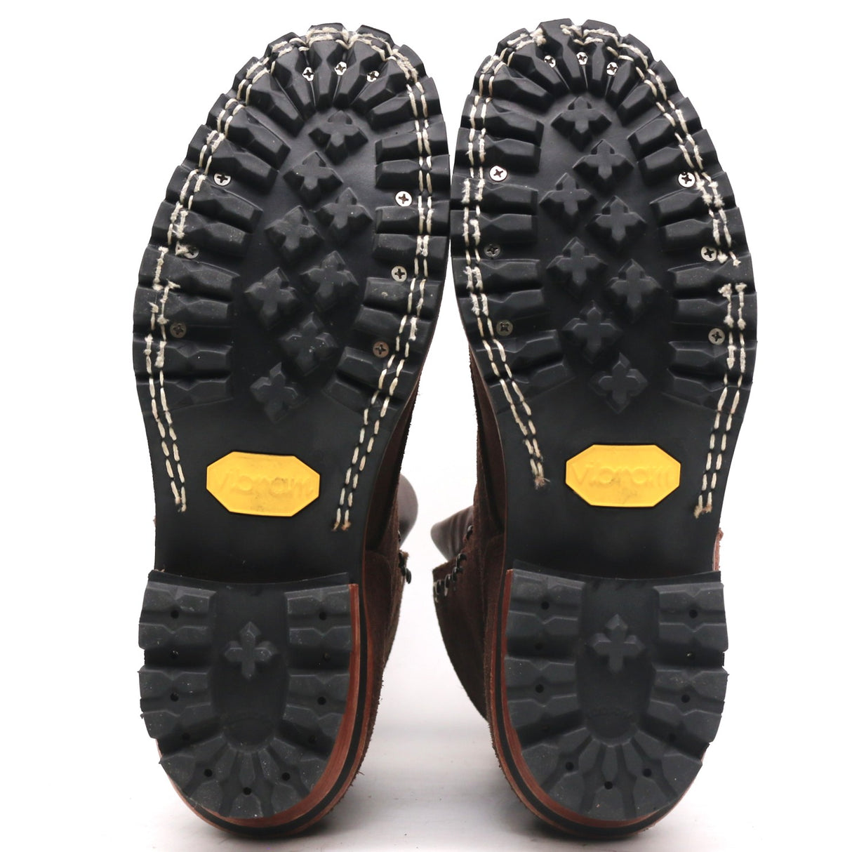 A pair of boots shown from the sole view, featuring a black Vibram treaded outsole with a distinctive yellow Vibram logo on the heel and white contrast stitching along the edge.