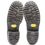 A pair of black heavy-duty boots shown from the sole, displaying deep lugs, stitching, and yellow Vibram logos on a white background.