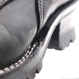 Close-up of a black leather boot corner showing a worn, scuffed edge between the leather vamp and the black lug sole, with white stitching along the edge.