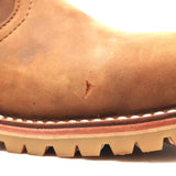 Close-up of a tan leather boot showing the toe area, a small scratch on the side, white stitching, and a chunky lug sole on a white background.