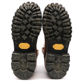 The soles of a pair of brown boots, showing deep lugs, Vibram branding in yellow on the heel, and a rugged black tread pattern on a white background.