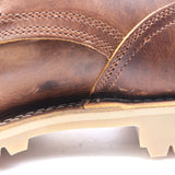 Close-up of a brown leather boot showing the textured leather upper, contrast stitching, and a light beige sole edge against a white background.