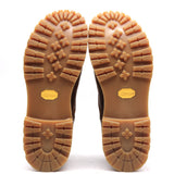 A pair of boots shown from the bottom, displaying their tan Vibram rubber soles with deep tread patterns and yellow Vibram logos on a white background.