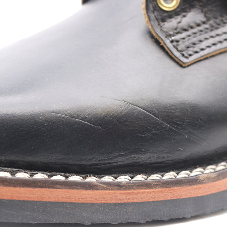 Close-up of a black leather boot toe showing scuffed leather texture, white stitching, a brown midsole layer, and part of a brown pull tab with a brass grommet.