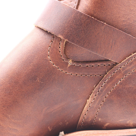 Close-up of a brown leather boot showing the texture, stitching, and strap details against a white background.