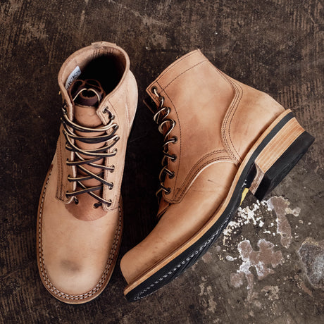 A pair of tan leather lace-up ankle boots with dark soles, arranged on a dark textured rustic surface.