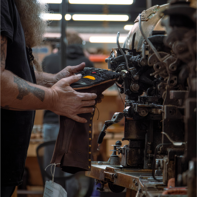A person in a workshop holds a dark brown leather boot while it is being assembled or repaired on a large industrial sewing or stitching machine. The workshop has warm overhead lighting and blurred background figures.