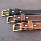 3 belts with brass hardware sitting on a metal table. One belt is black, one belt is dark brown, and one belt is medium brown.