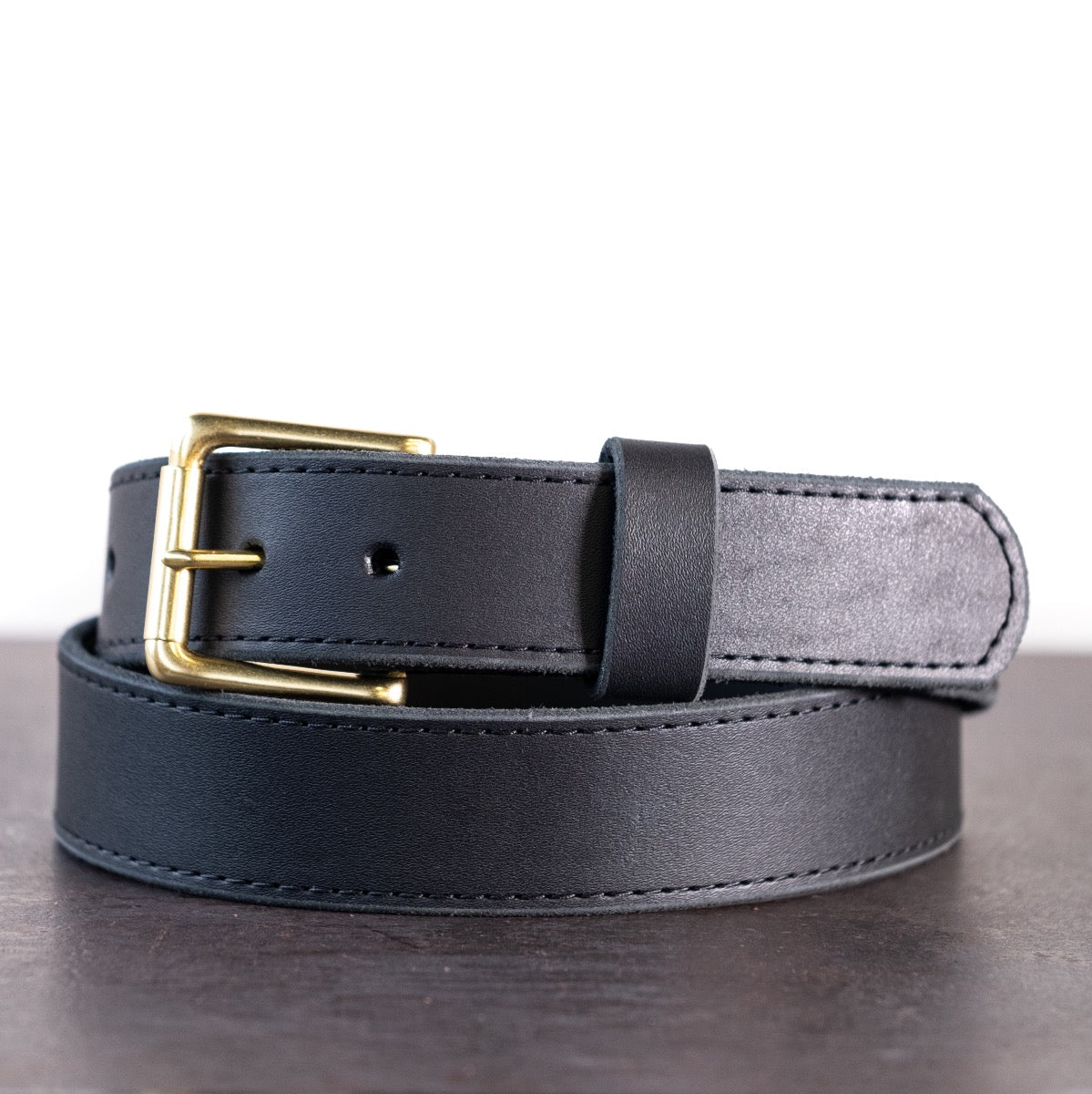 Brown belt with brown stitching and brass hardware sitting on metal surface with white background