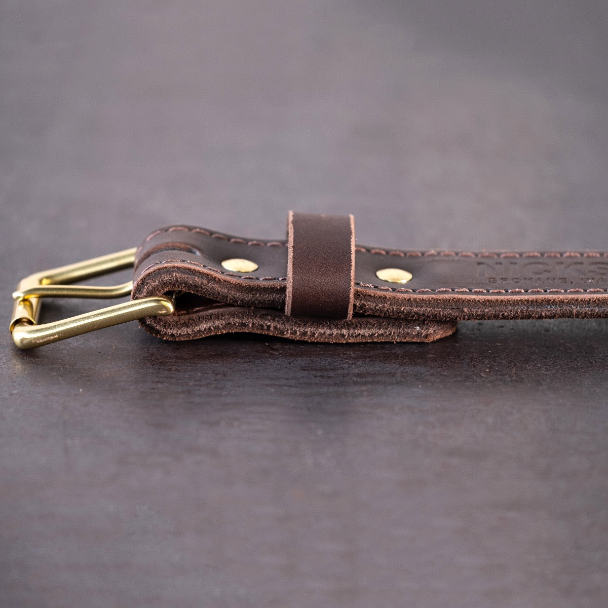 Dark brown belt with brown stitching and brass hardware sitting on metal surface