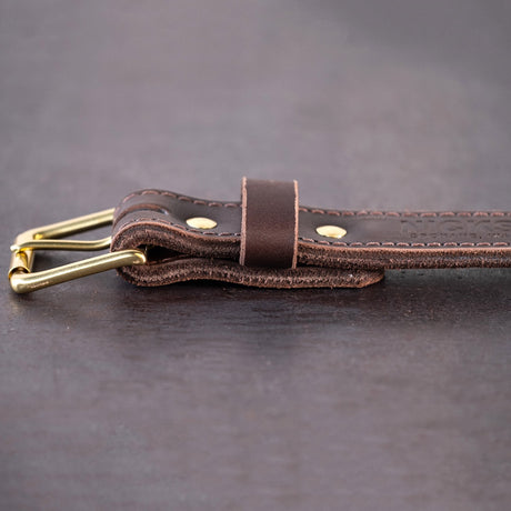 Dark brown belt with brown stitching and brass hardware sitting on metal surface