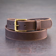 Dark brown belt with brown stitching and brass hardware sitting on metal surface