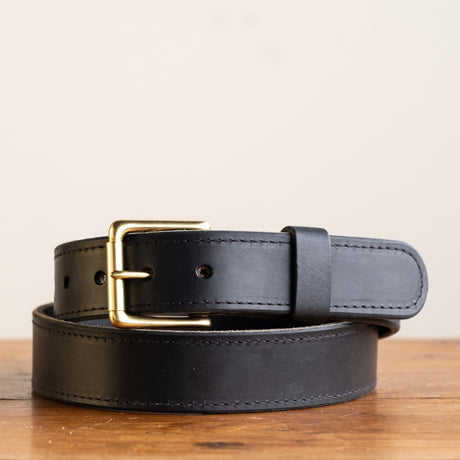 Close-up of a black leather belt with a smooth finish, featuring a simple brass buckle and visible grain details, showcasing its timeless and rustic craftsmanship.