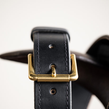 Close-up of a black leather belt with a smooth finish, featuring a simple brass buckle and visible grain details, showcasing its timeless and rustic craftsmanship.