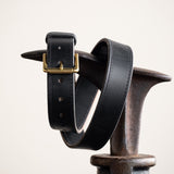 Close-up of a black leather belt with a smooth finish, featuring a simple brass buckle and visible grain details, showcasing its timeless and rustic craftsmanship.