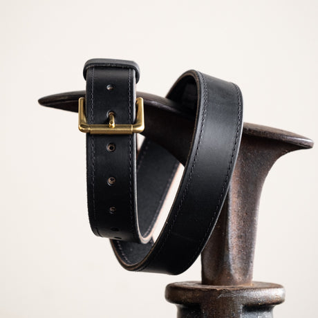 Close-up of a black leather belt with a smooth finish, featuring a simple brass buckle and visible grain details, showcasing its timeless and rustic craftsmanship.