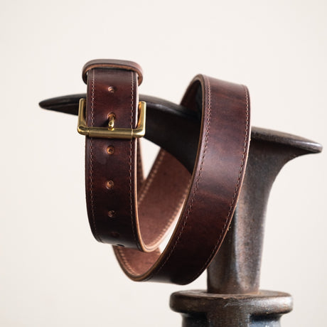 Close-up of a brown leather belt with a smooth finish, featuring a simple brass buckle and visible grain details, showcasing its timeless and rustic craftsmanship.